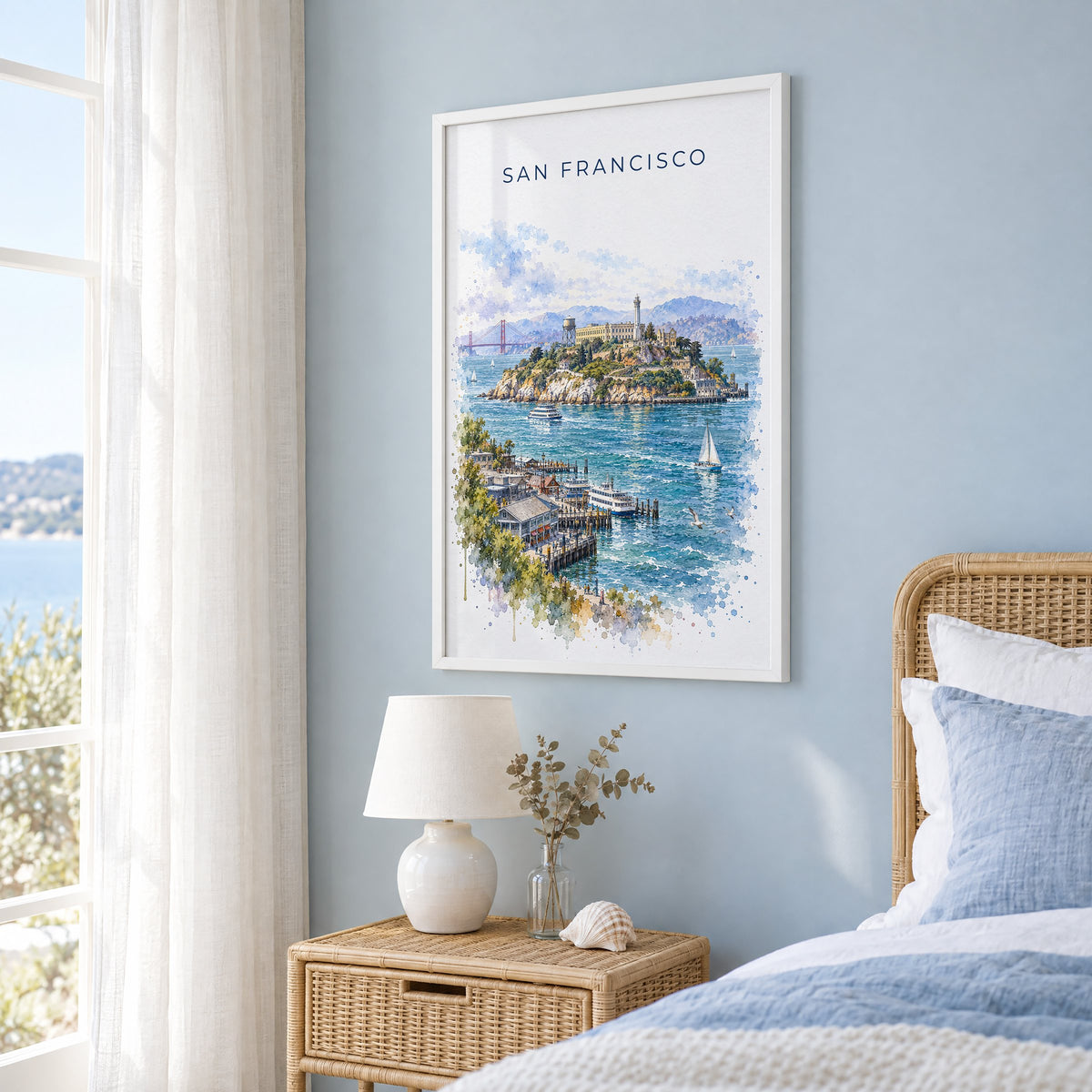 Alcatraz Island watercolor print in a white frame on a pale blue wall beside a rattan bed in a coastal bedroom