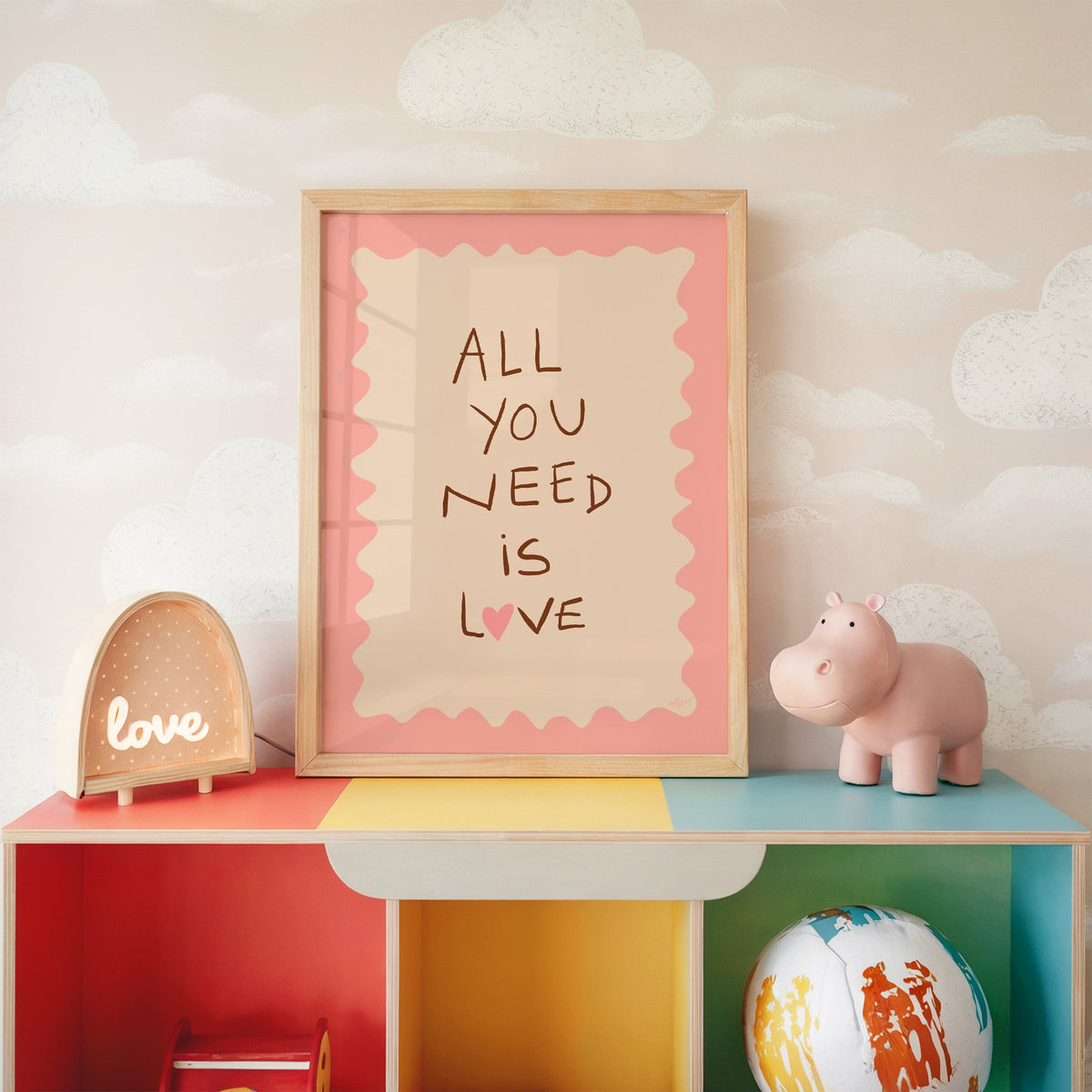 All you need is love lettering nursery print in an oak frame on a rainbow toy shelf against cloud wallpaper