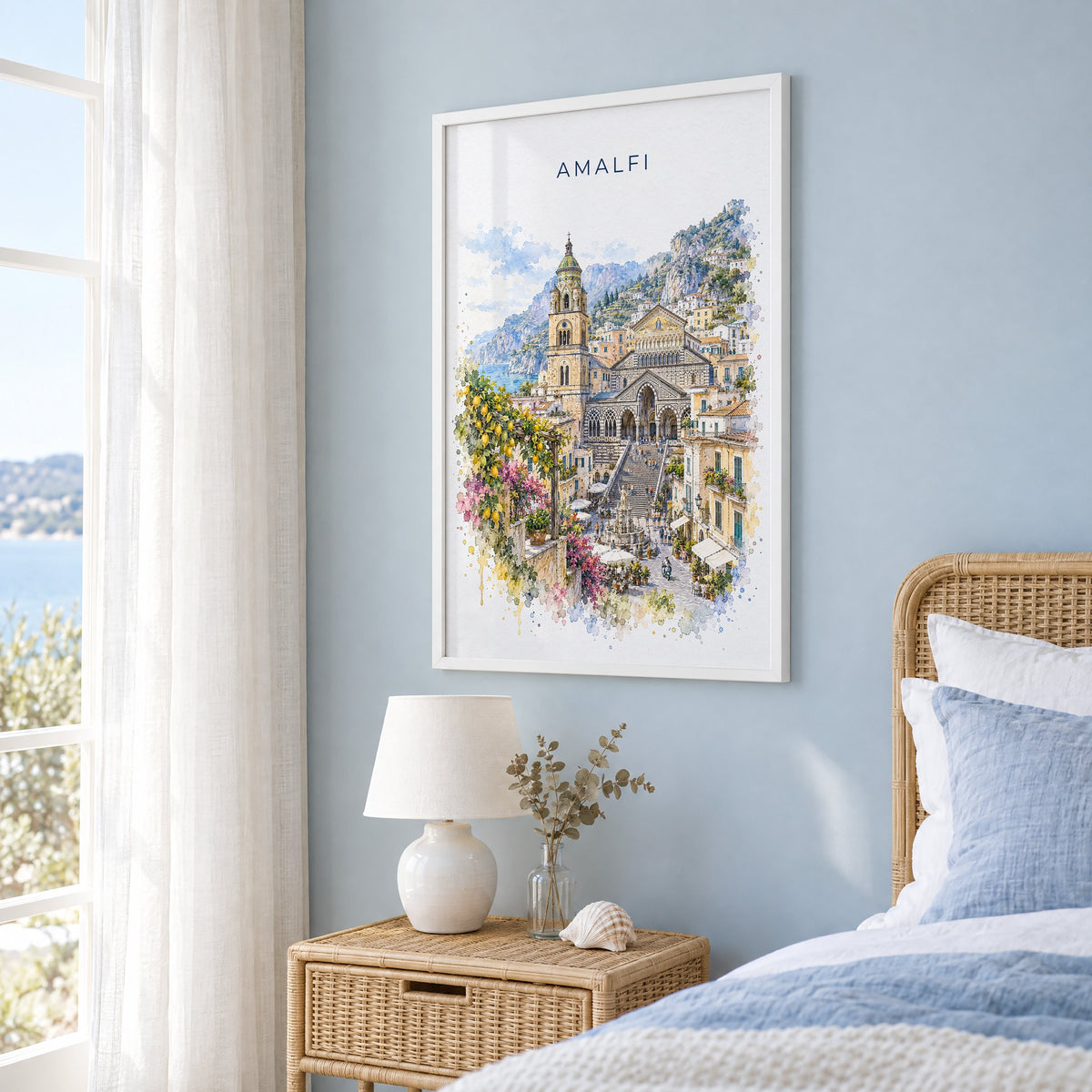 Amalfi watercolor print in a white frame on a pale blue wall beside a rattan bed in a coastal bedroom