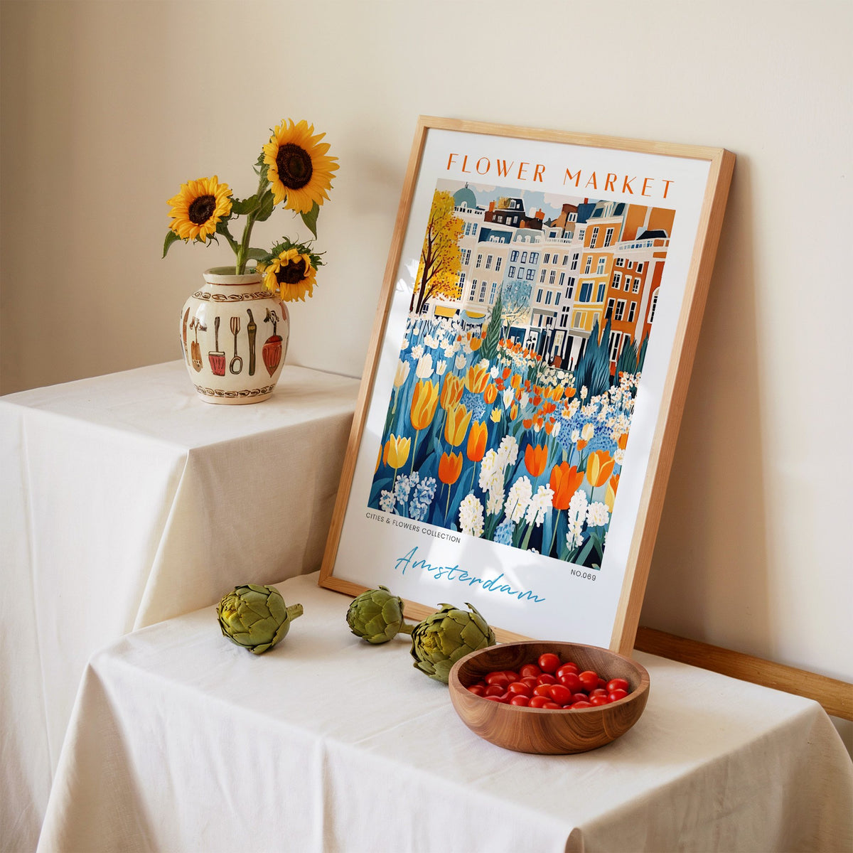 Amsterdam flower market print in an oak frame leaning on a linen-draped table beside a vase of sunflowers