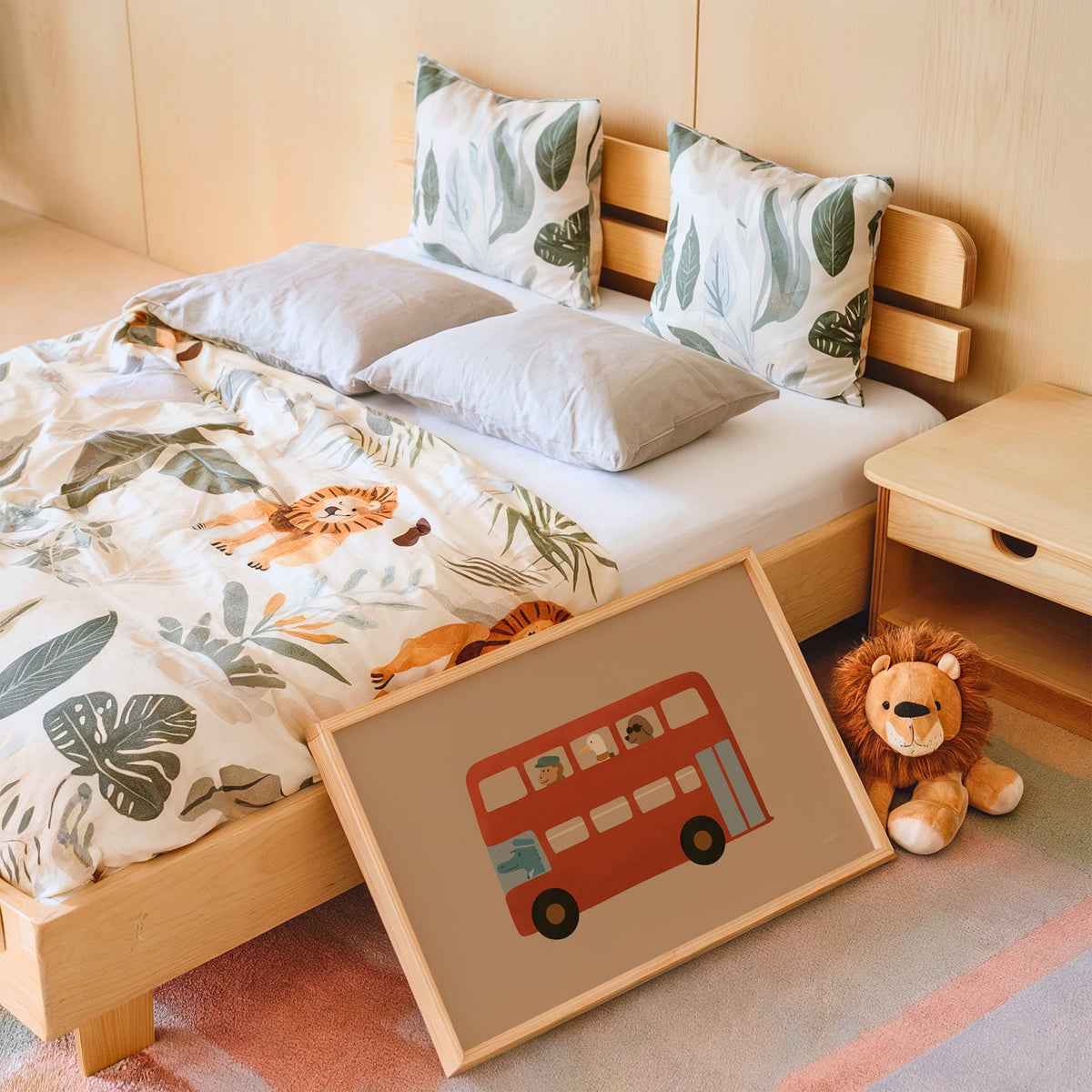 Animals riding red double decker bus nursery print in an oak frame leaning on a kids bed by a lion plush