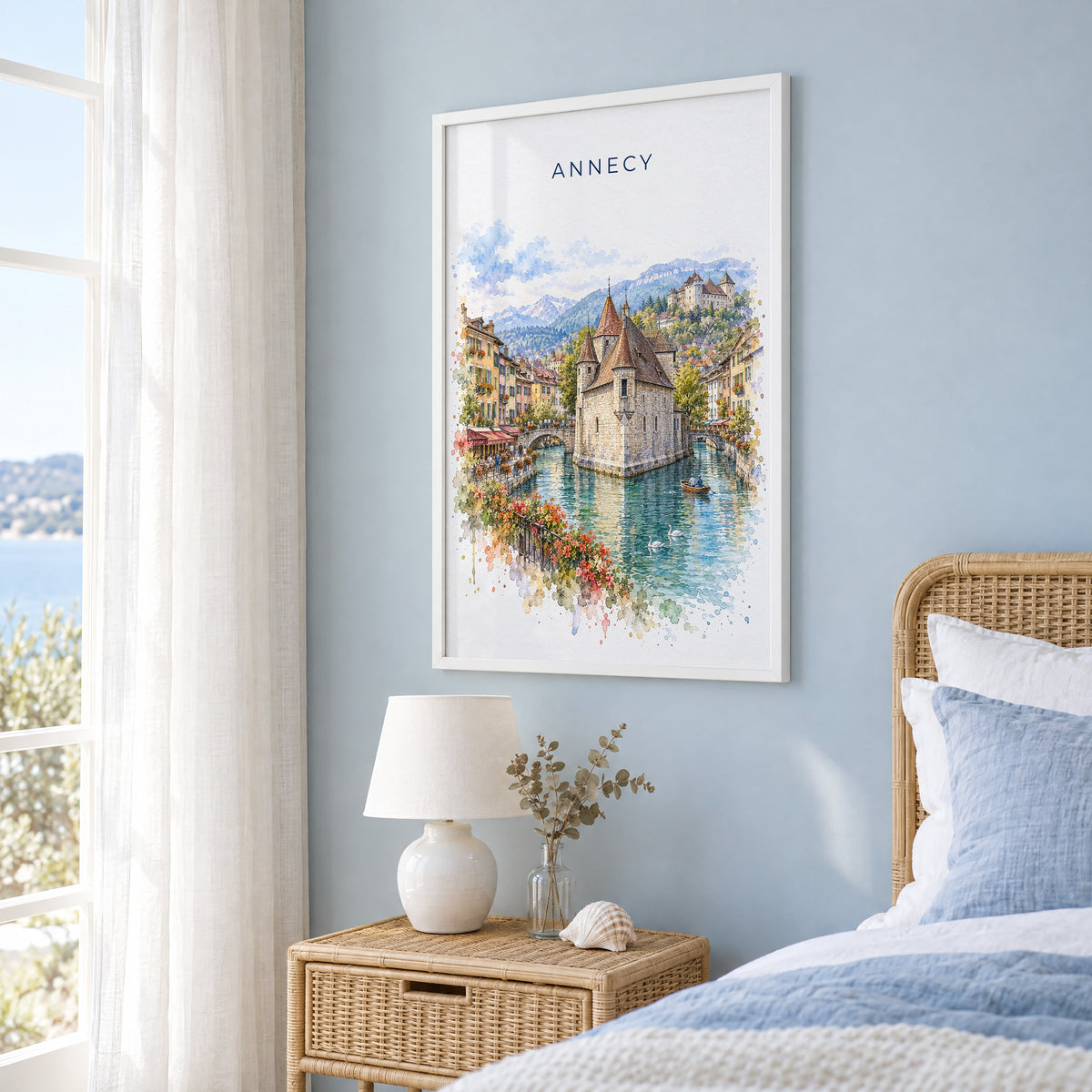 Annecy watercolor print in a white frame on a pale blue wall beside a rattan bed in a coastal bedroom