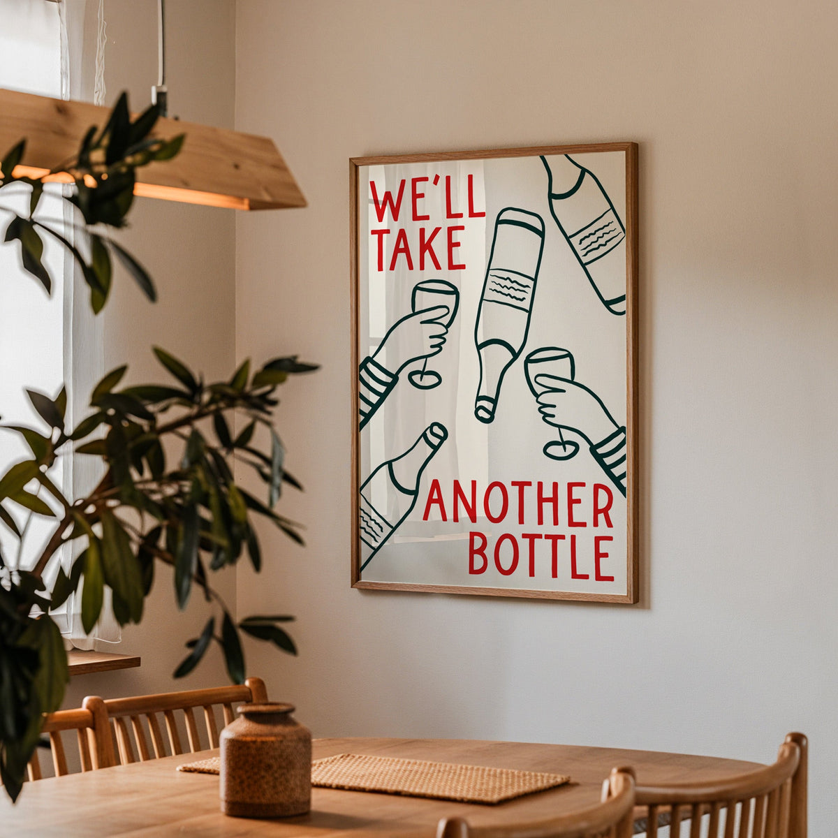 We'll Take Another Bottle wine print in a walnut frame above a dining table beside an olive tree