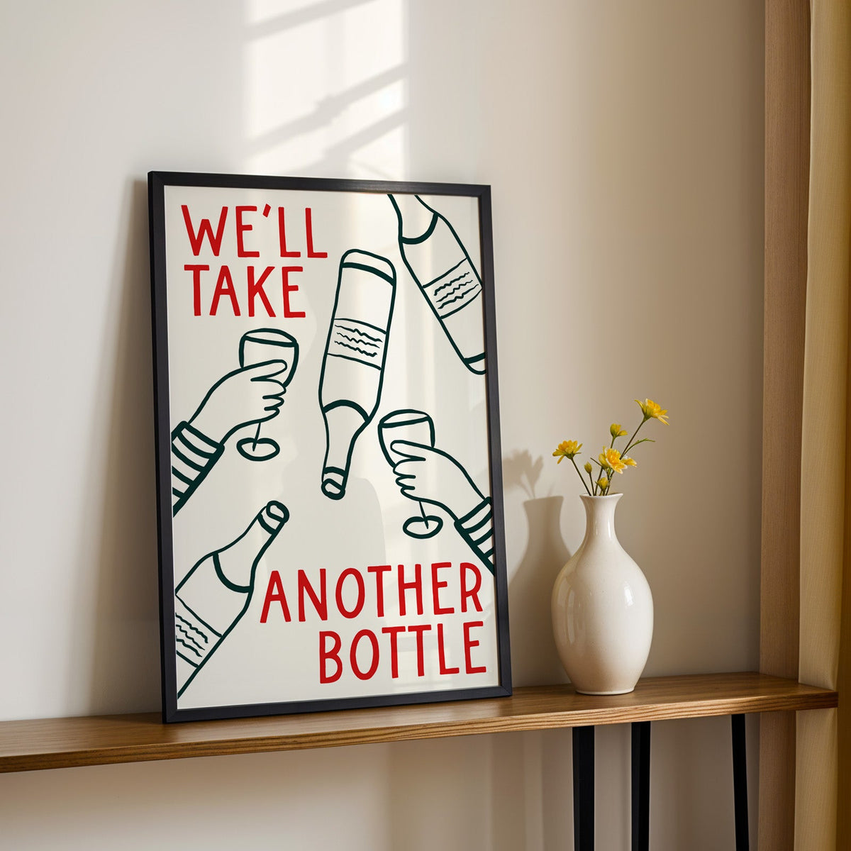 We'll Take Another Bottle wine print in a black frame on a console table with yellow daisies