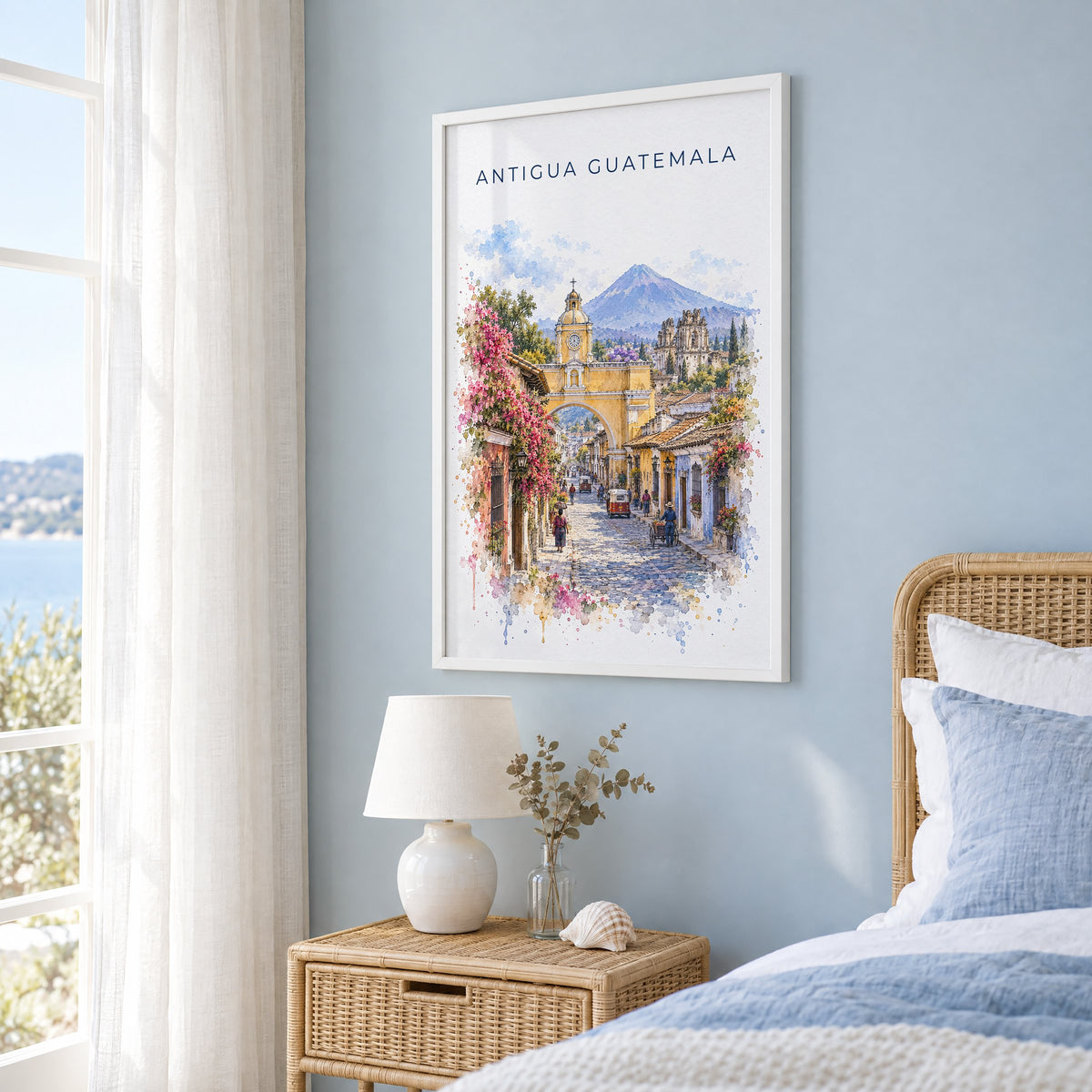 Antigua Guatemala watercolor print in a white frame on a pale blue wall beside a rattan bed in a coastal bedroom