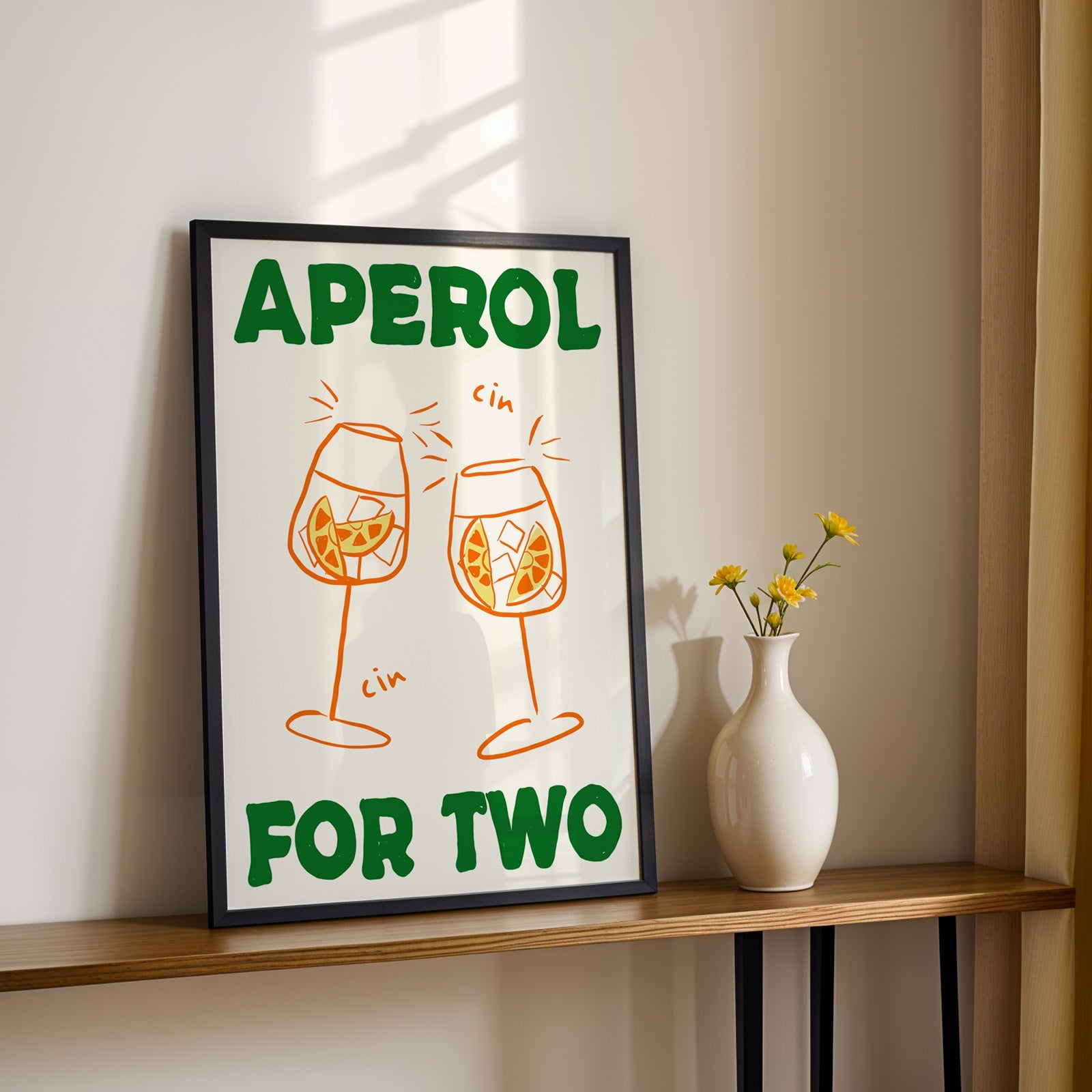 Aperol spritz print in a black frame on a console table with yellow daisies