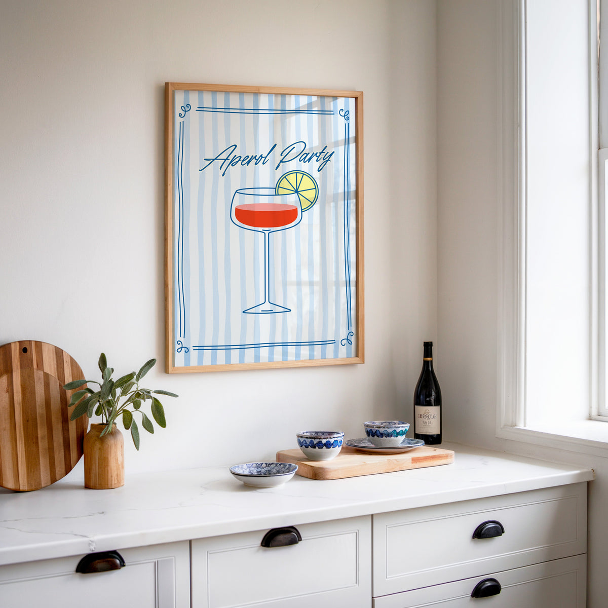 Aperol Party print in an oak frame above a white sideboard with a bottle of wine