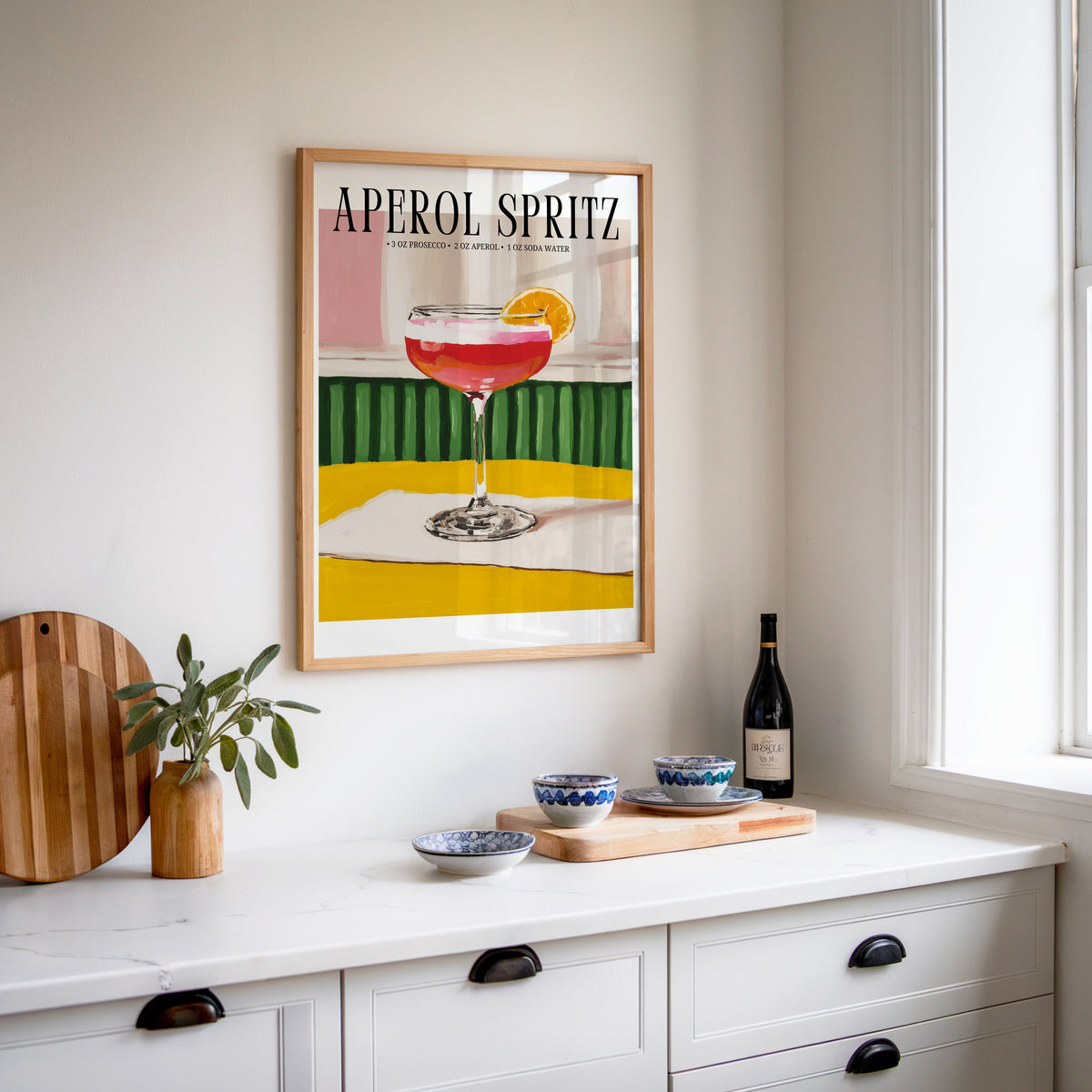 Aperol Spritz recipe print in an oak frame above a white sideboard with a bottle of wine