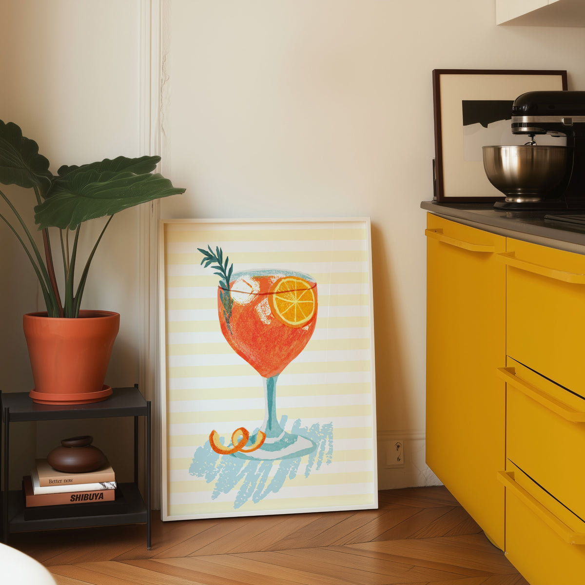 Aperol spritz print in a white frame leaning on parquet beside a bright yellow kitchen