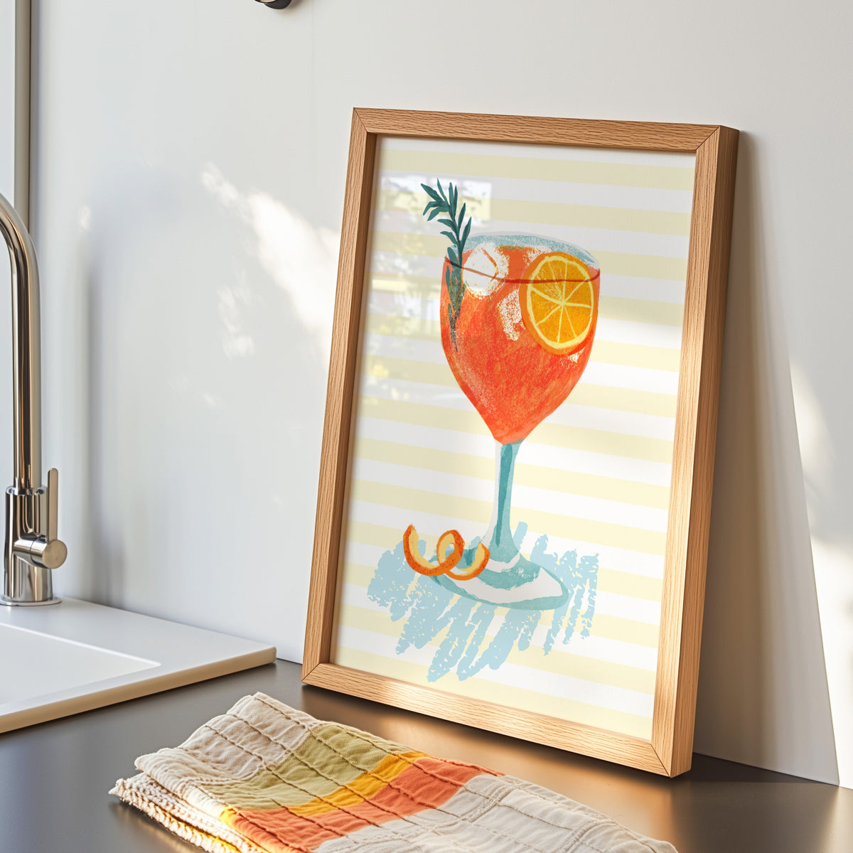 Aperol spritz print in an oak frame by the kitchen sink beside a striped tea towel