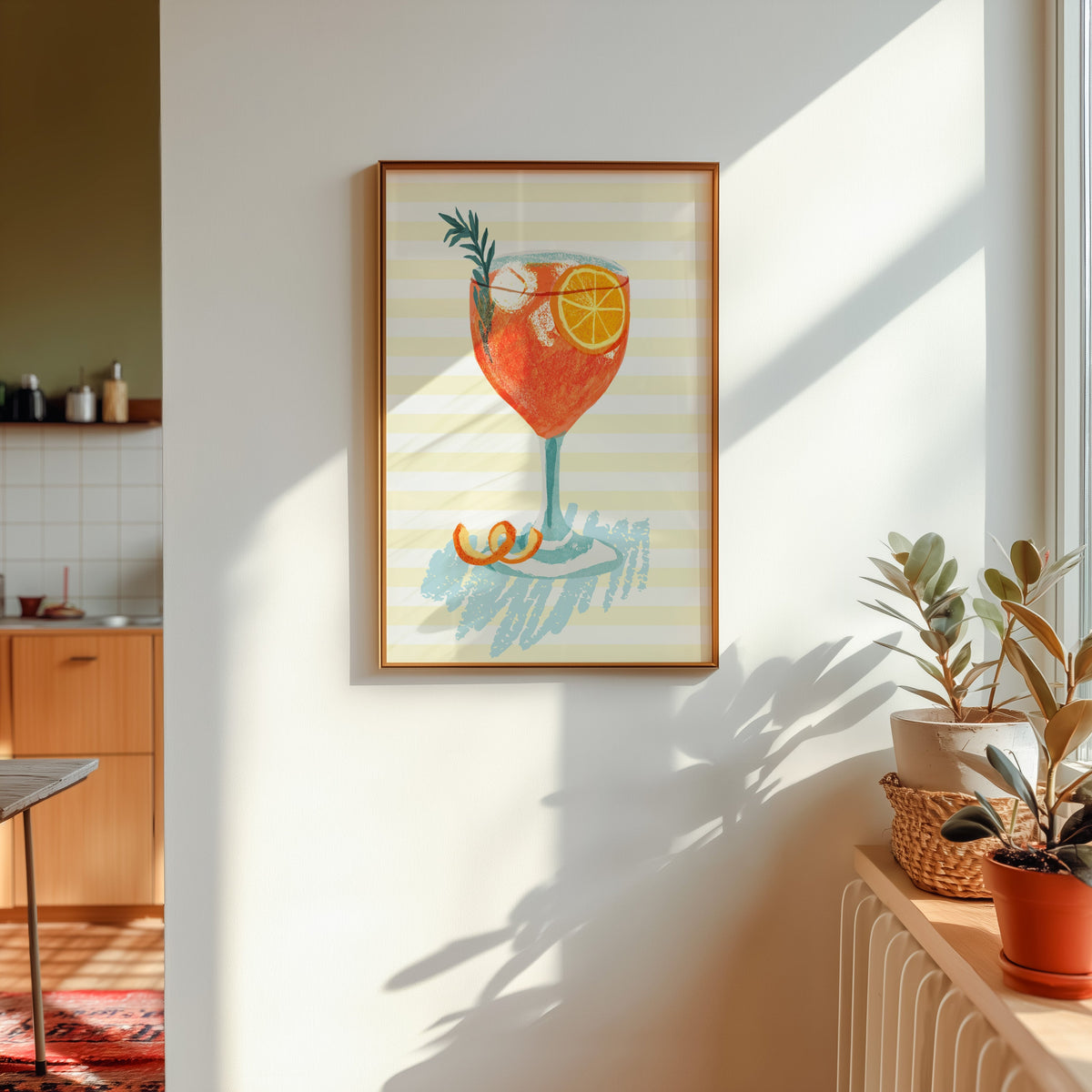 Aperol spritz print shown in a gold frame in sunlight with plants and a kitchen beyond