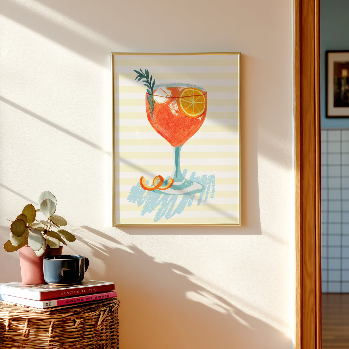 Aperol spritz print shown in a gold frame above a basket of books and a pilea plant