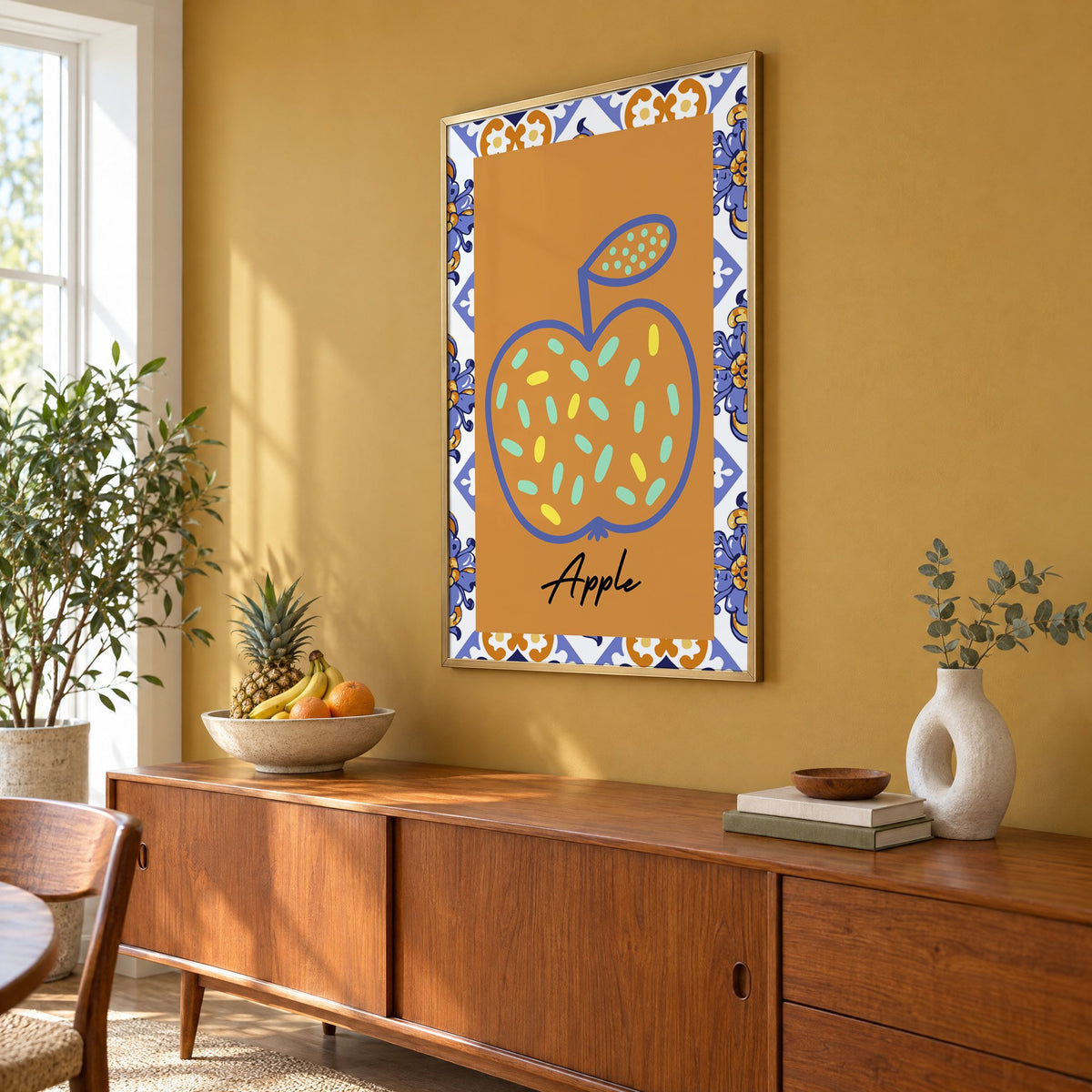 Apple blue outline ochre majolica print in a brass frame on a mustard wall above a teak sideboard with a fruit bowl, seen at an angle