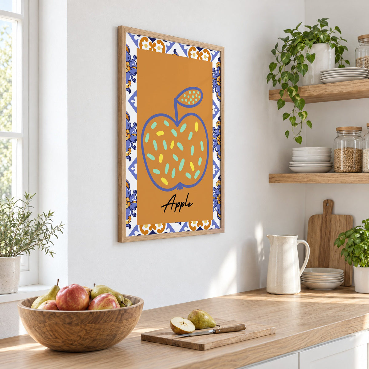 Apple blue outline ochre majolica print in an oak frame on a white kitchen wall above a counter with apples and pears, seen at an angle