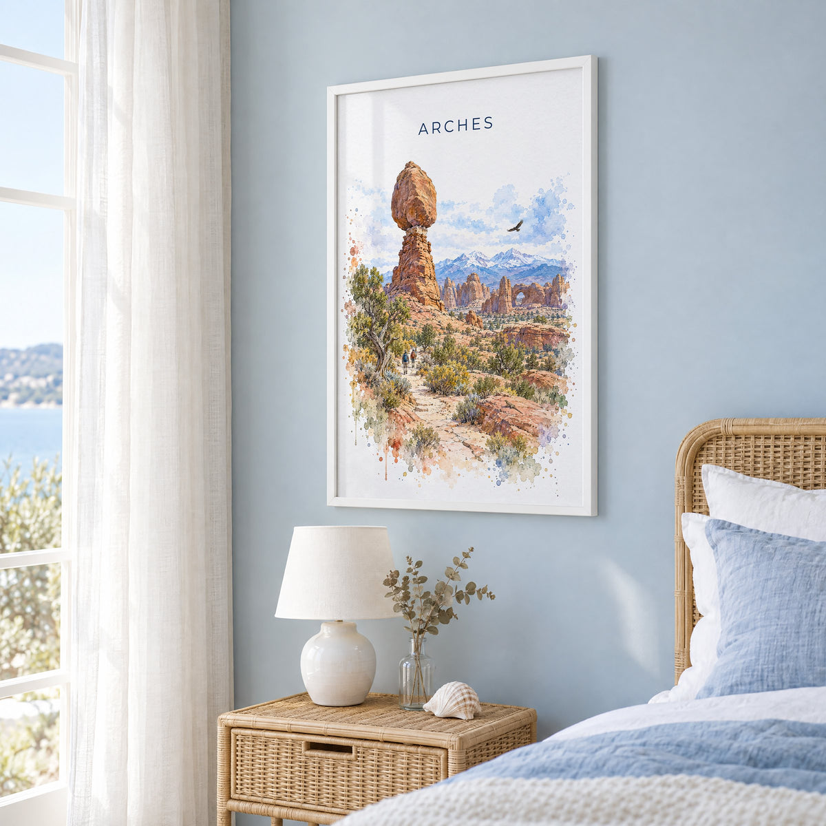 Arches National Park watercolor print in a white frame on a pale blue wall beside a rattan bed in a coastal bedroom