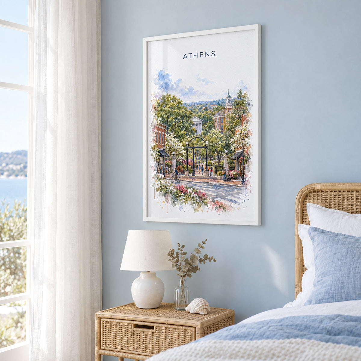 Athens, Georgia watercolor print in a white frame on a pale blue wall beside a rattan bed in a coastal bedroom