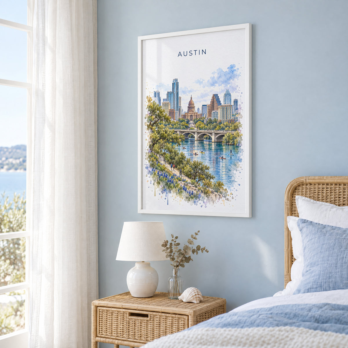 Austin watercolor print in a white frame on a pale blue wall beside a rattan bed in a coastal bedroom