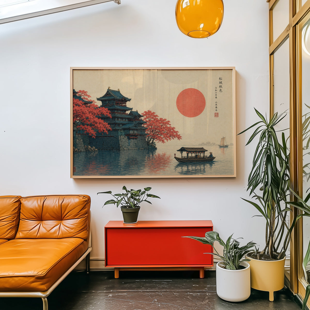 Autumn castle on a lake with red sun print in a light wood frame above a red cabinet beside a tan leather daybed