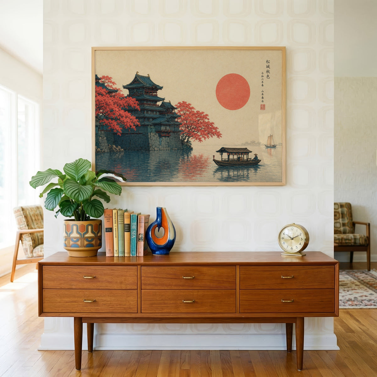 Autumn castle on a lake with red sun print in a light oak frame above a teak credenza with books and a brass clock