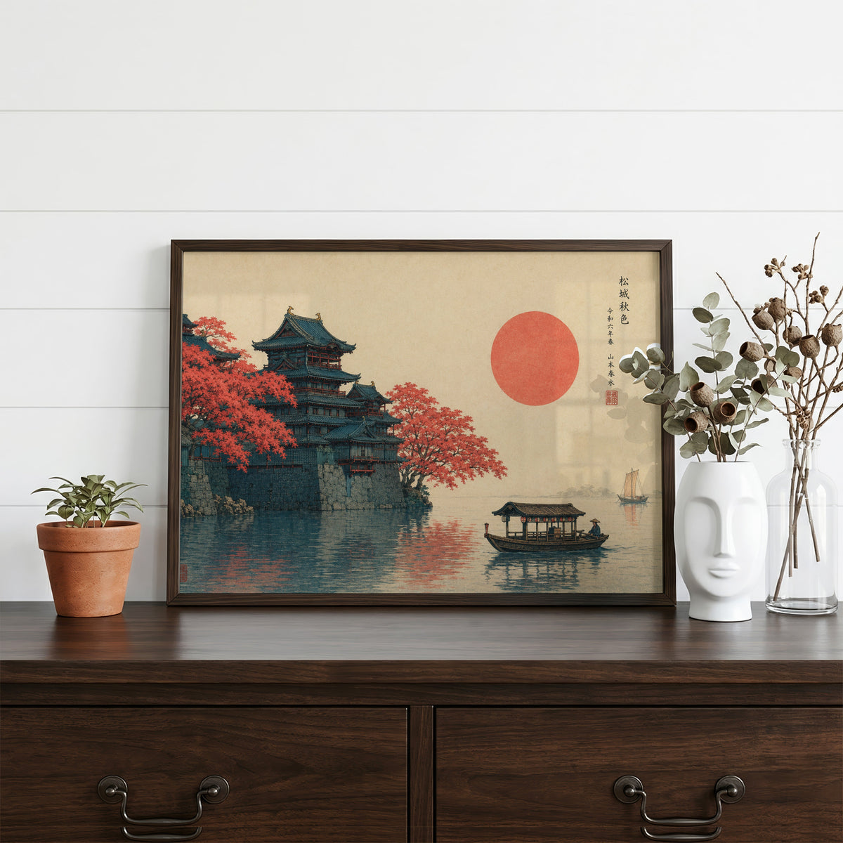Autumn castle on a lake with red sun print in a dark walnut frame on a dark wood dresser beside a white face vase