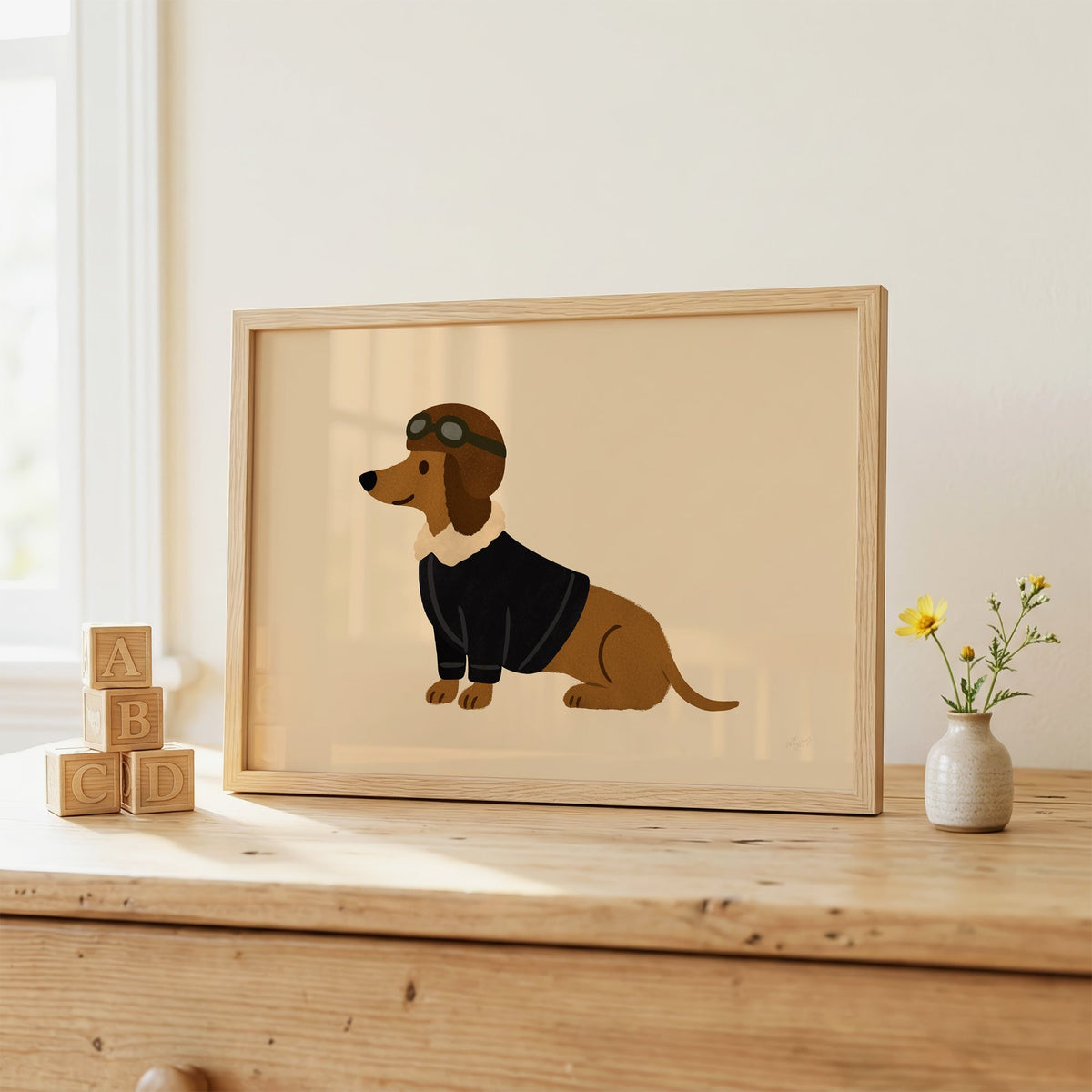 Dachshund in aviator cap nursery print in an oak frame on a wooden dresser beside ABC blocks