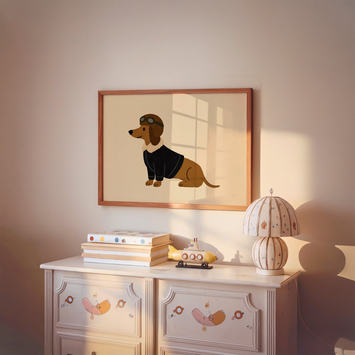 Dachshund in aviator cap nursery print in a cherry wood frame above a painted dresser and lamp