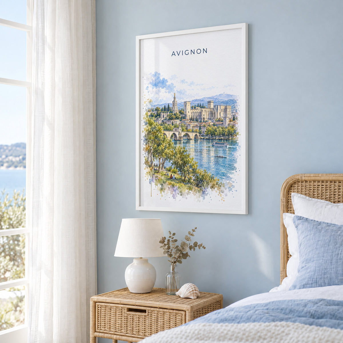 Avignon watercolor print in a white frame on a pale blue wall beside a rattan bed in a coastal bedroom