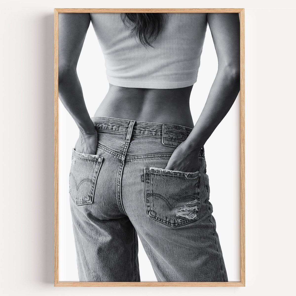 Hands in back pockets of distressed jeans print in a light oak frame on a plain white wall