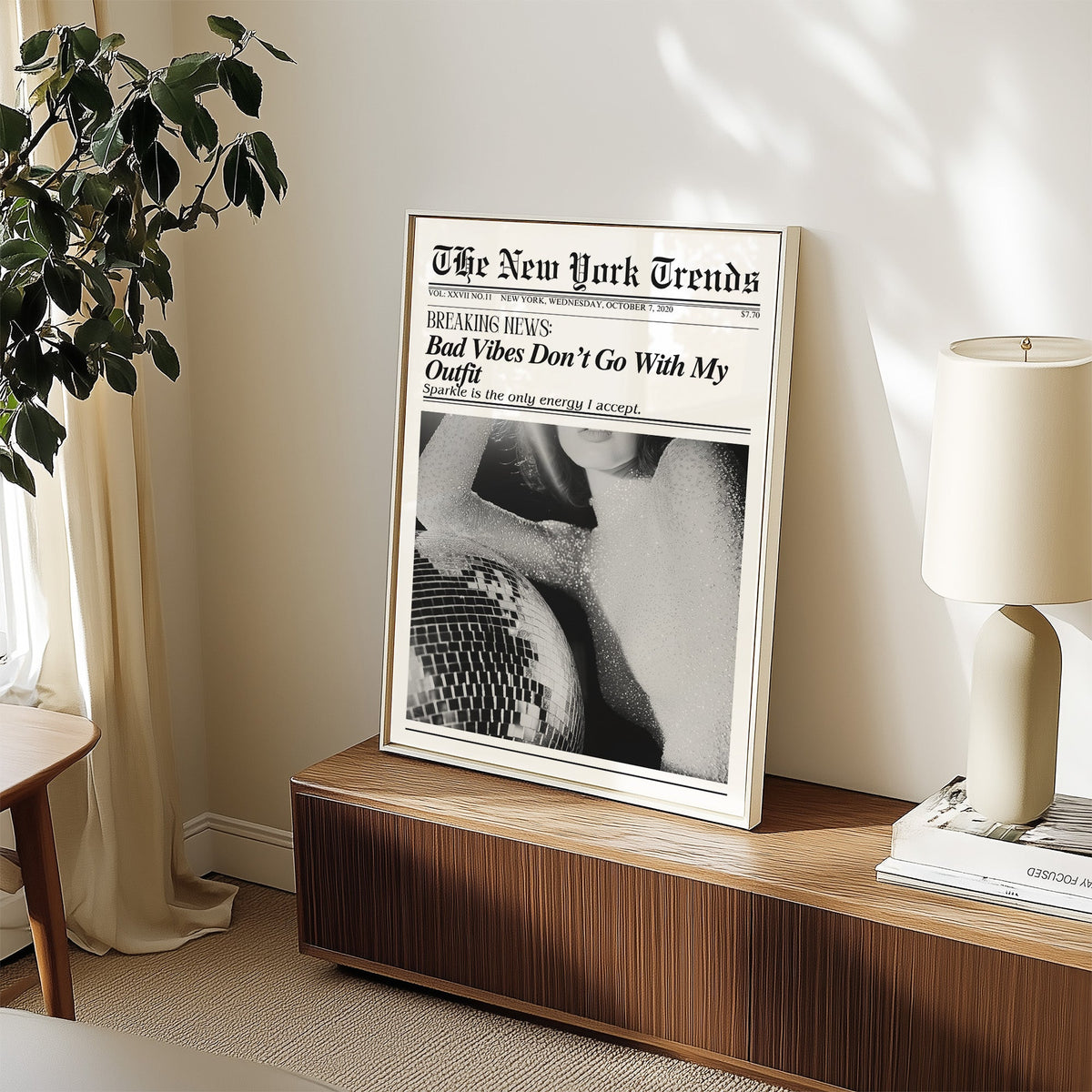 Sequin top and disco ball mock newspaper print in a white frame on a walnut sideboard beside a ceramic lamp