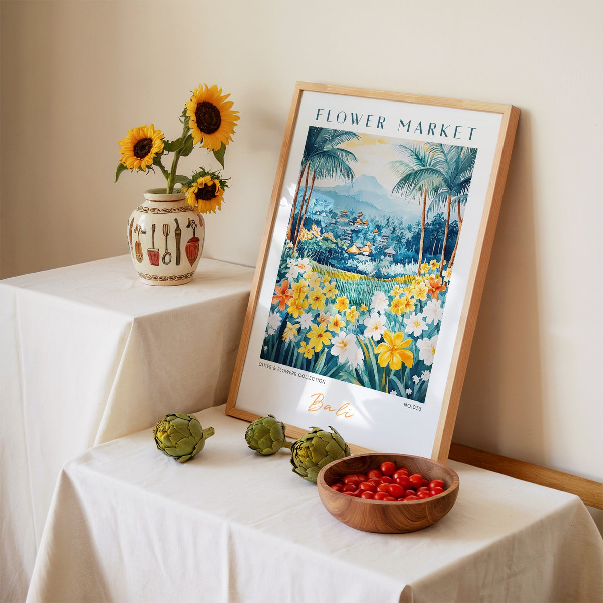 Bali flower market print in an oak frame leaning on a linen-draped table beside a vase of sunflowers
