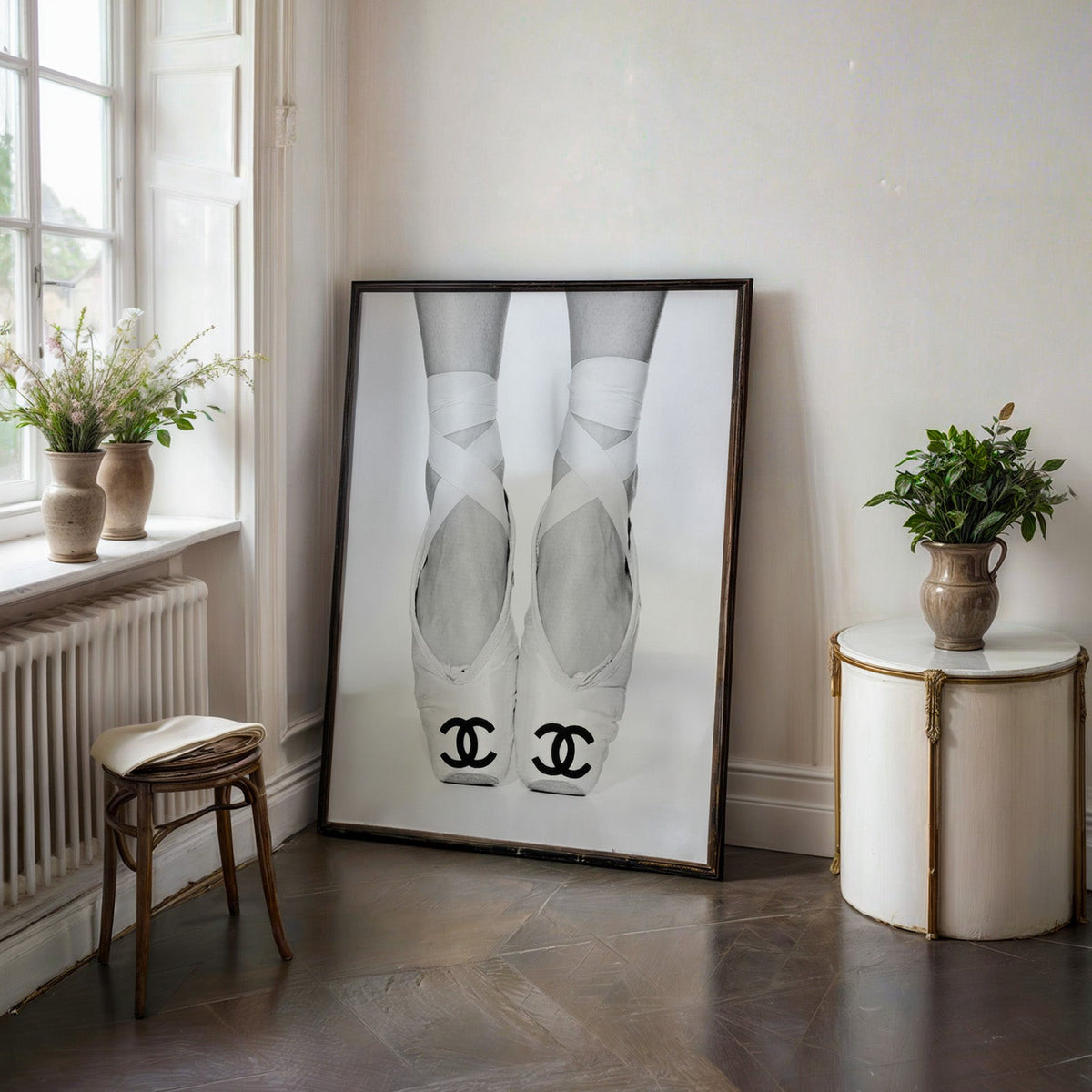 White ballet pointe shoes with crossed ribbons print in a black frame on a dark stone floor beside a white side table