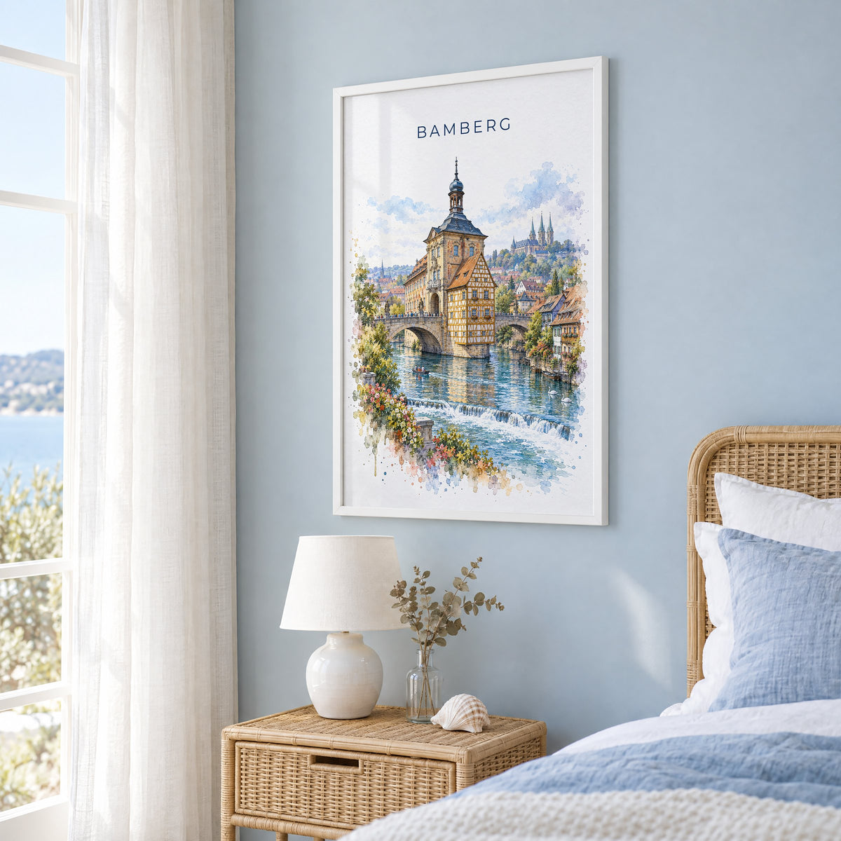 Bamberg watercolor print in a white frame on a pale blue wall beside a rattan bed in a coastal bedroom