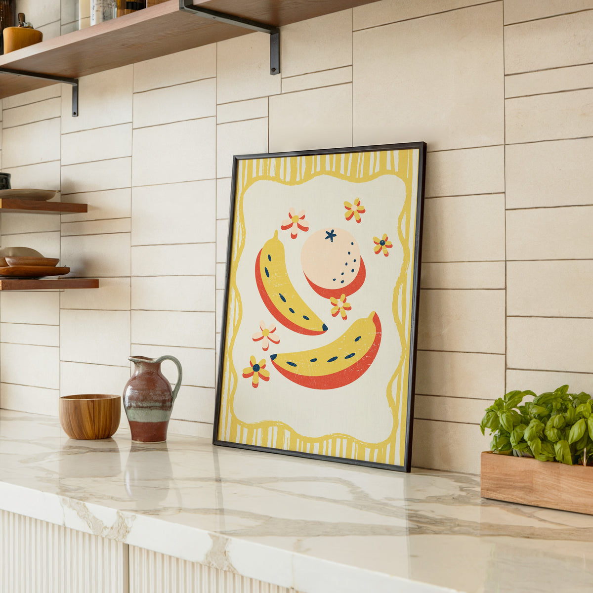 Banana and peach print in a black frame leaning on a marble counter in a tiled kitchen