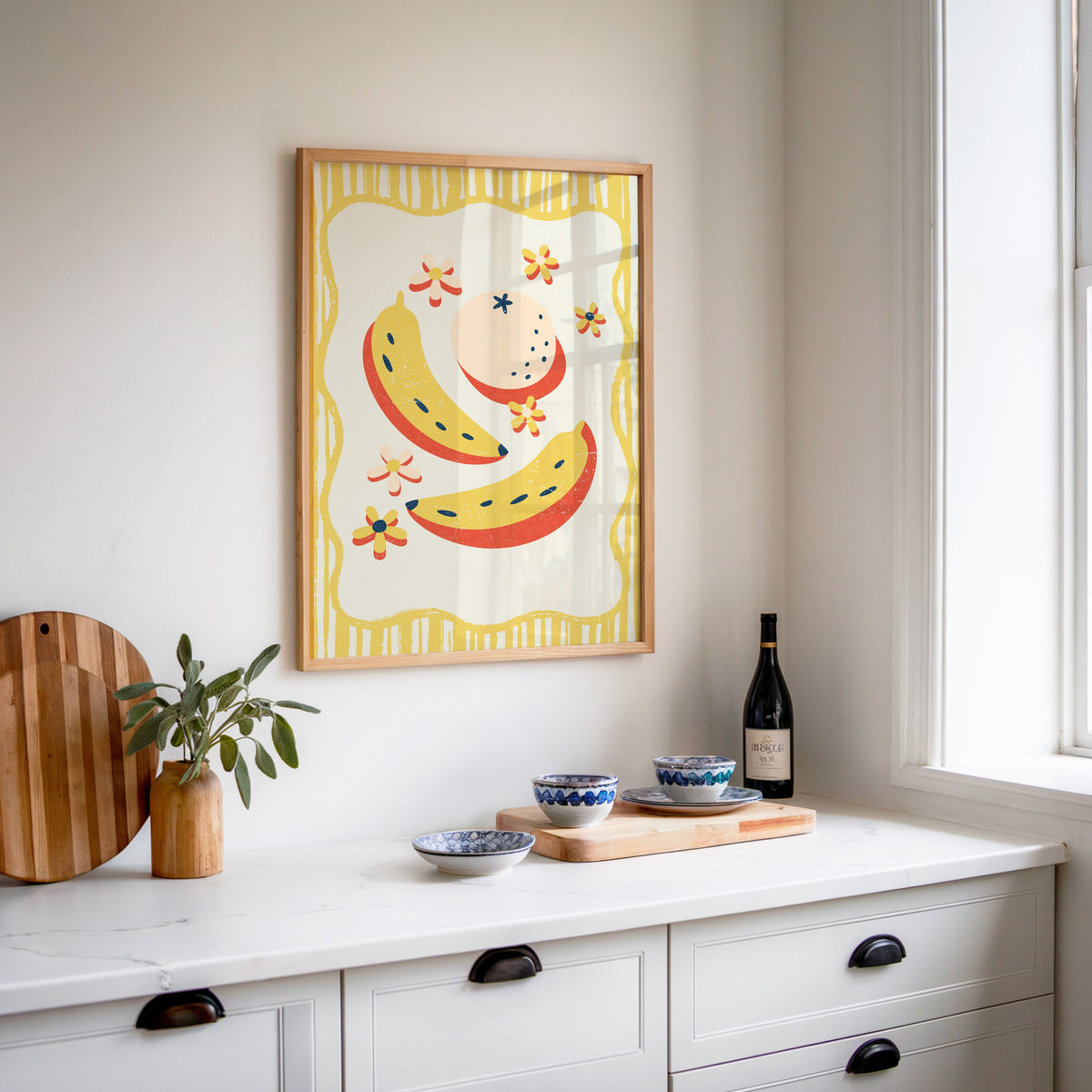 Banana and peach print in an oak frame above a white sideboard with a bottle of wine