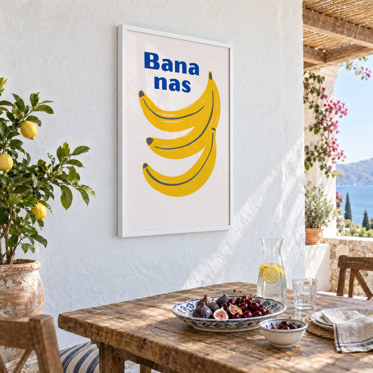 Bana nas blue type banana bunch print in a white frame on a whitewashed terrace wall above a lunch table with figs and cherries, seen at an angle