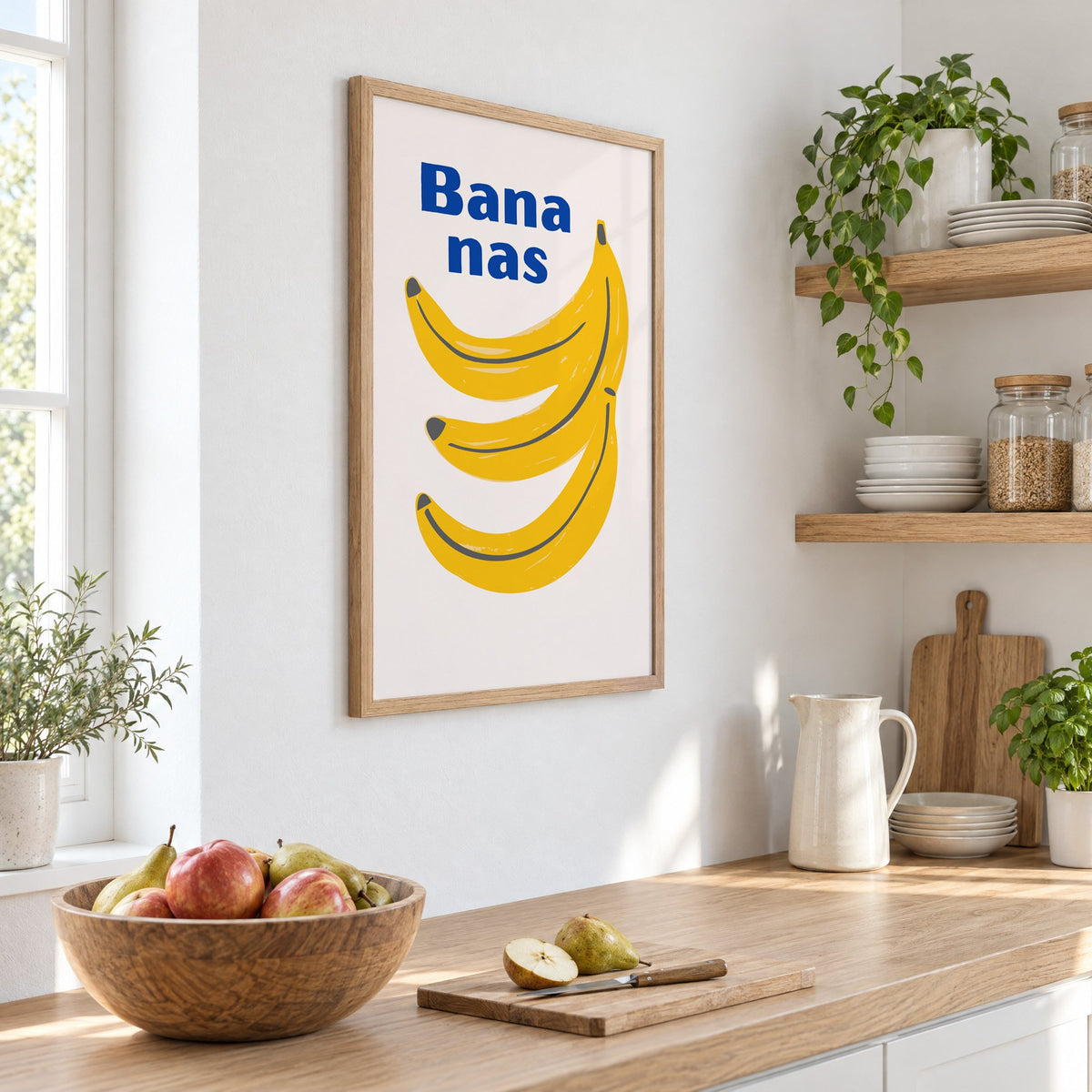 Bana nas blue type banana bunch print in an oak frame on a white kitchen wall above a counter with apples and pears, seen at an angle