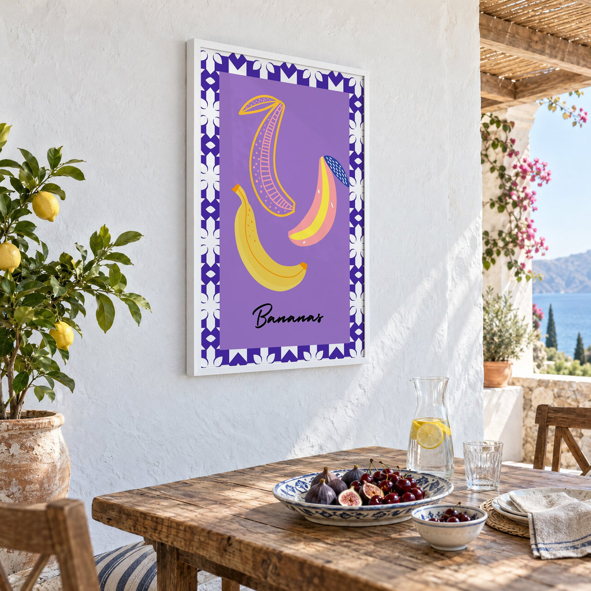 Bananas doodle lavender tile border print in a white frame on a whitewashed terrace wall above a lunch table with figs and cherries, seen at an angle