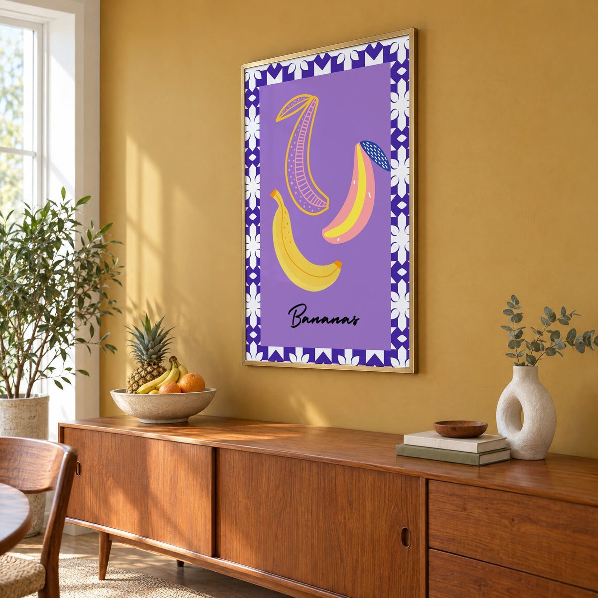 Bananas doodle lavender tile border print in a brass frame on a mustard wall above a teak sideboard with a fruit bowl, seen at an angle