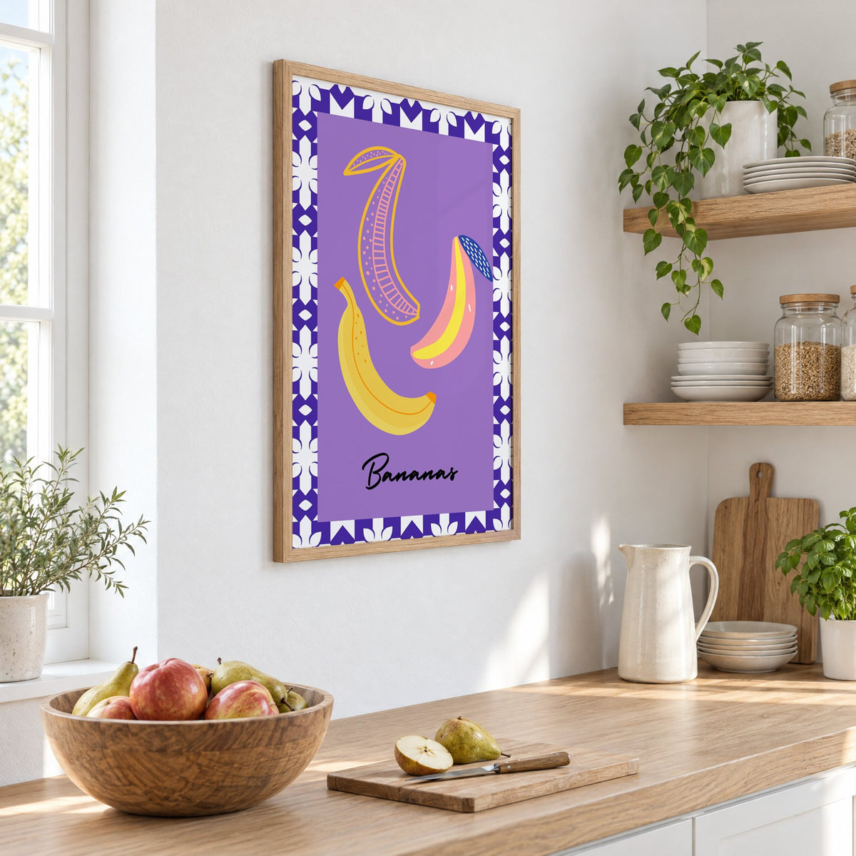 Bananas doodle lavender tile border print in an oak frame on a white kitchen wall above a counter with apples and pears, seen at an angle