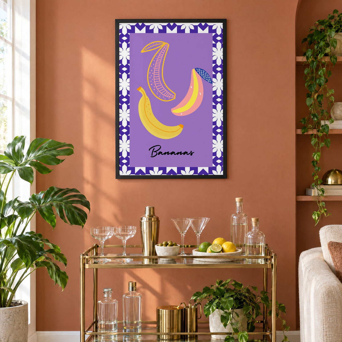 Bananas doodle lavender tile border print in a black frame on a terracotta wall above a brass bar cart with coupes and citrus