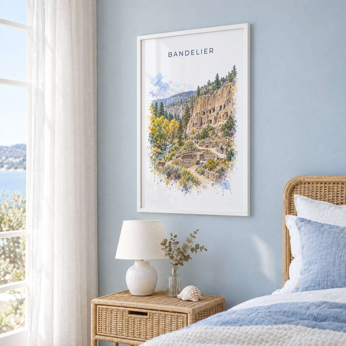 Bandelier National Monument watercolor print in a white frame on a pale blue wall beside a rattan bed in a coastal bedroom