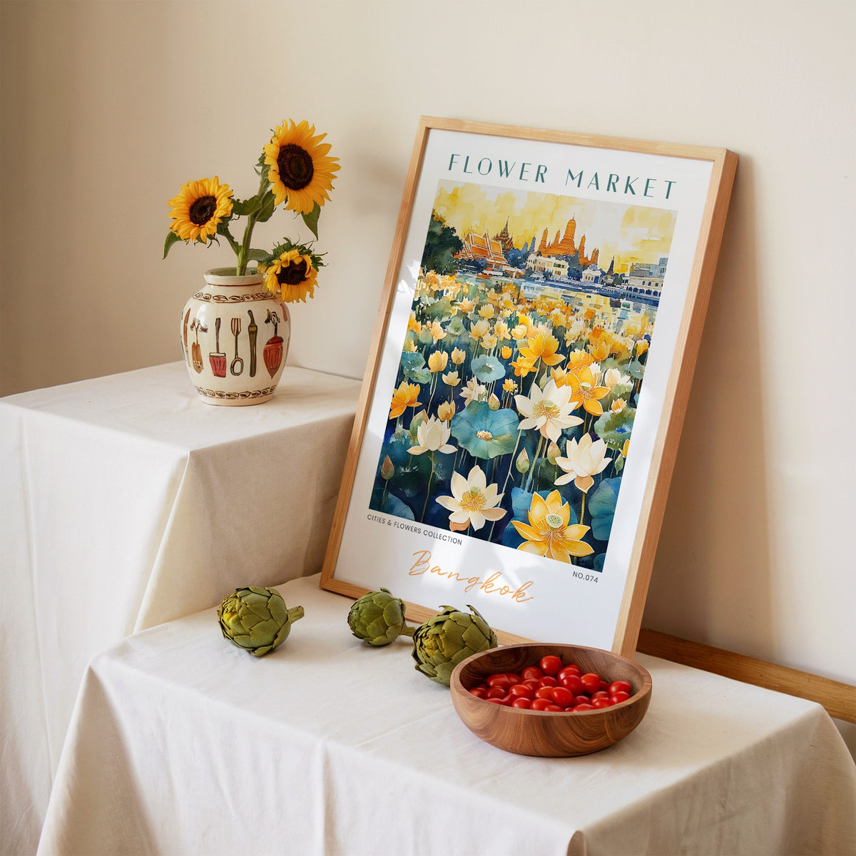 Bangkok flower market print in an oak frame leaning on a linen-draped table beside a vase of sunflowers