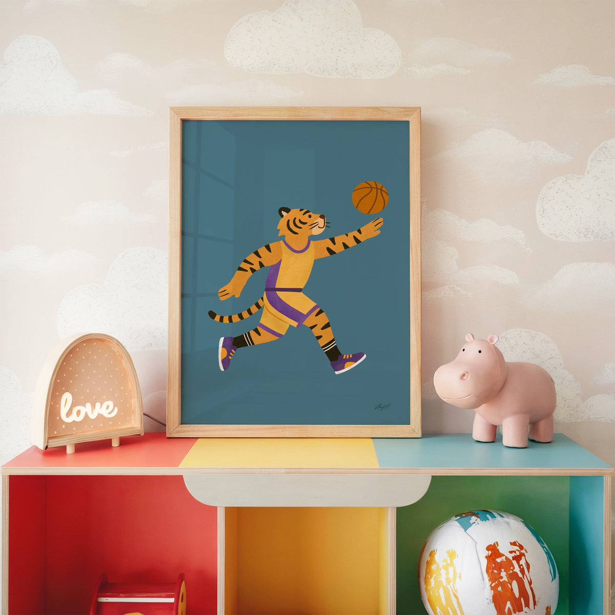 Tiger playing basketball nursery print in an oak frame on a rainbow toy shelf against cloud wallpaper