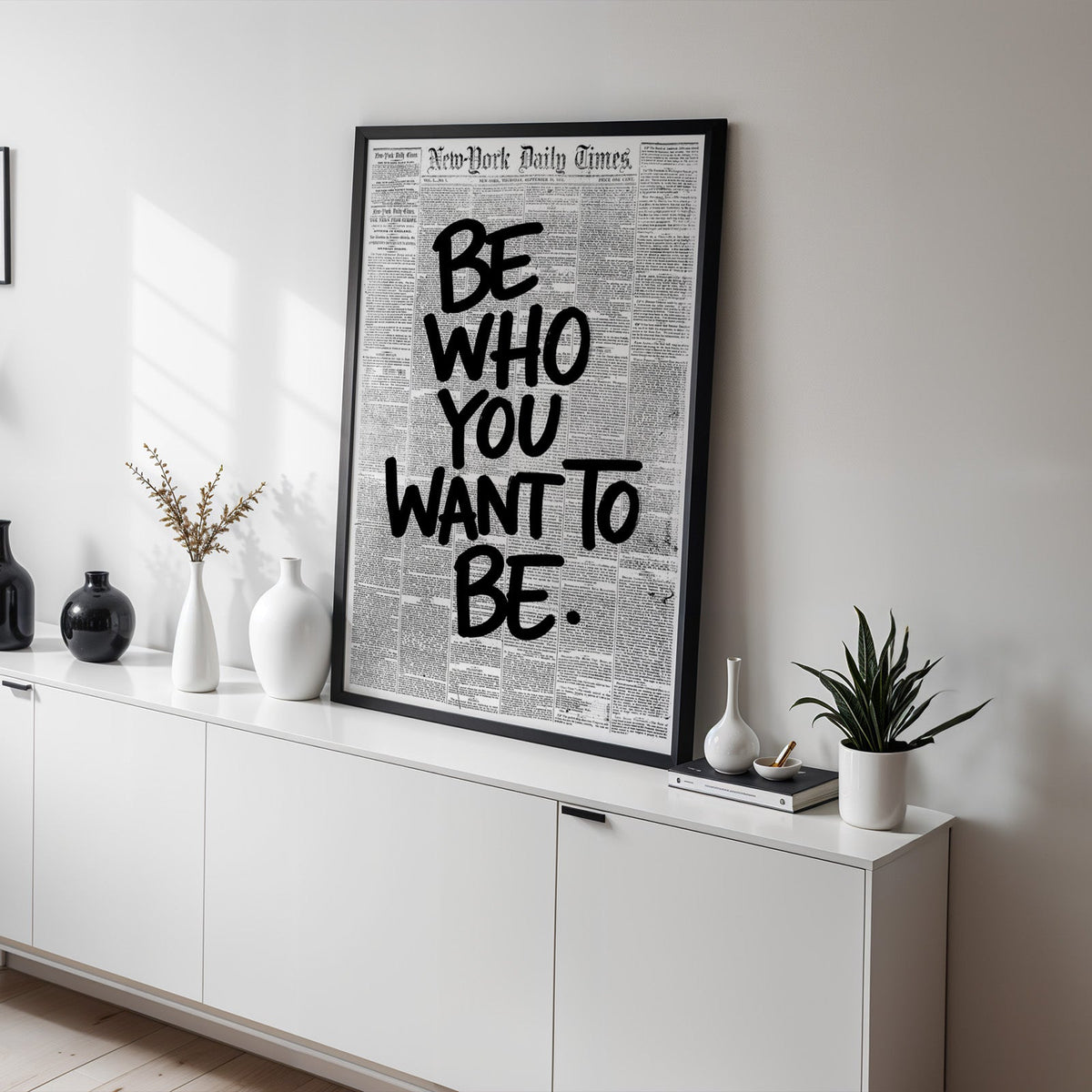 Brush lettering over vintage newspaper page print in a black frame on a white sideboard with black and white vases