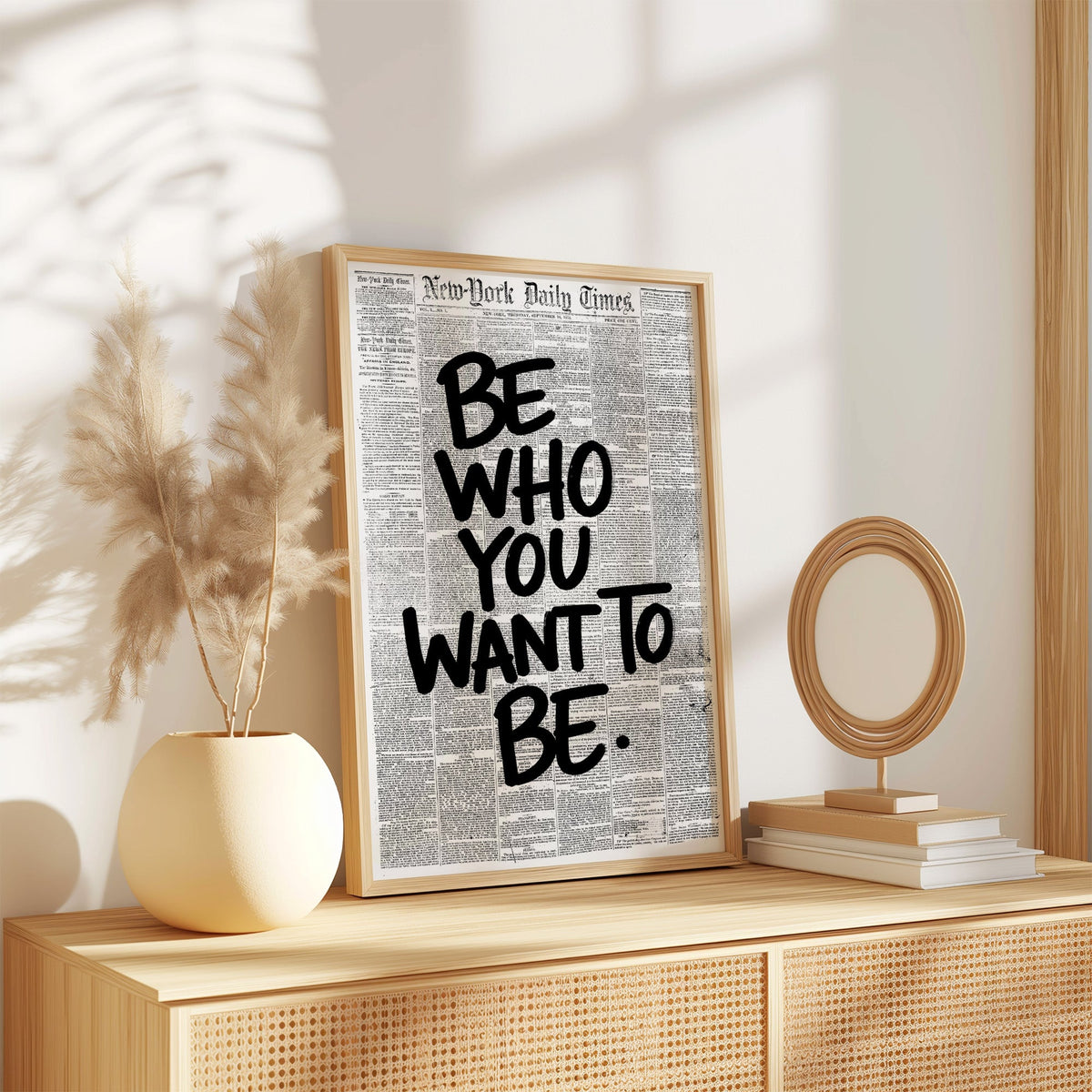 Brush lettering over vintage newspaper page print in an oak frame on a rattan sideboard beside pampas grass
