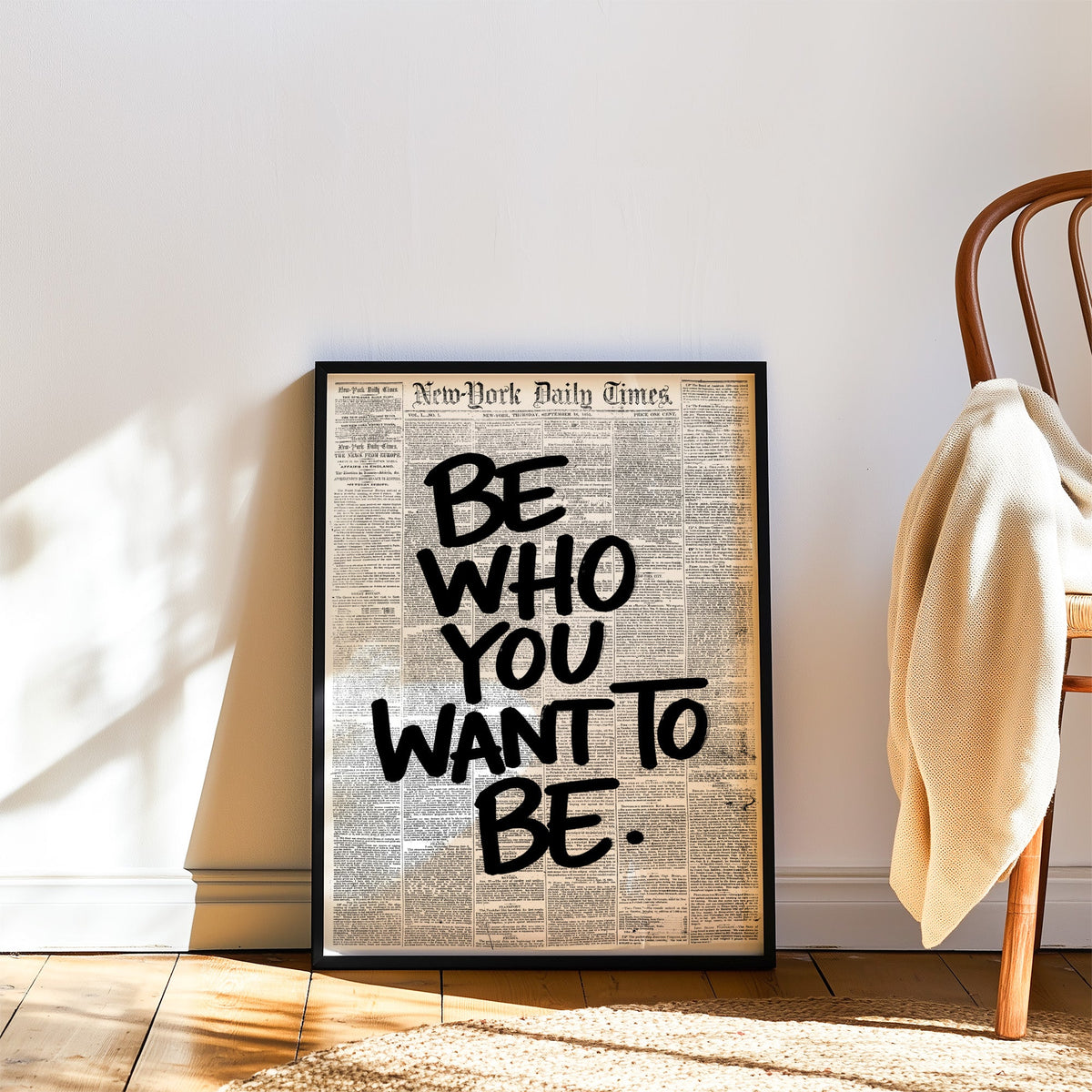 Brush lettering over vintage newspaper page print in a black frame leaning on a wood floor beside a bentwood chair