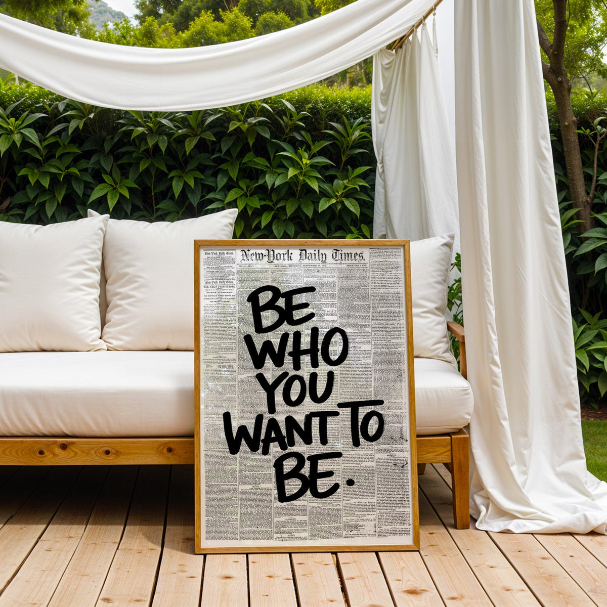 Brush lettering over vintage newspaper page print in an oak frame on an outdoor daybed under white drapes