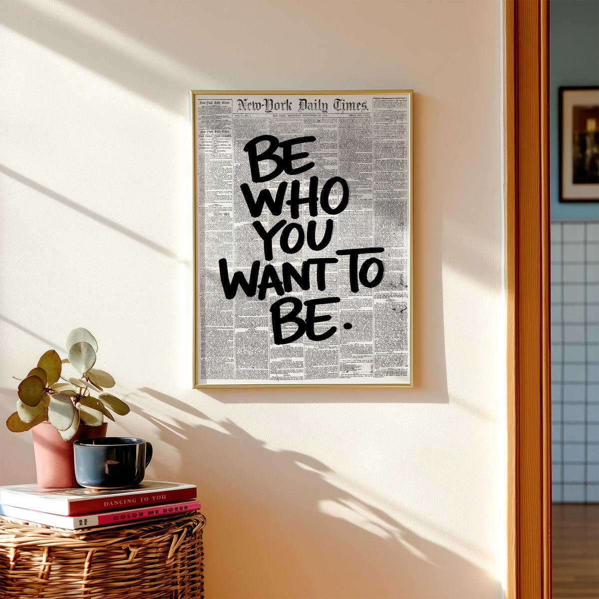 Brush lettering over vintage newspaper page print shown in a gold frame above a wicker basket and stacked books