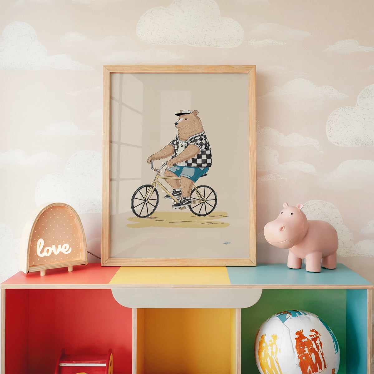 Brown bear riding yellow bicycle nursery print in an oak frame on a rainbow toy shelf against cloud wallpaper