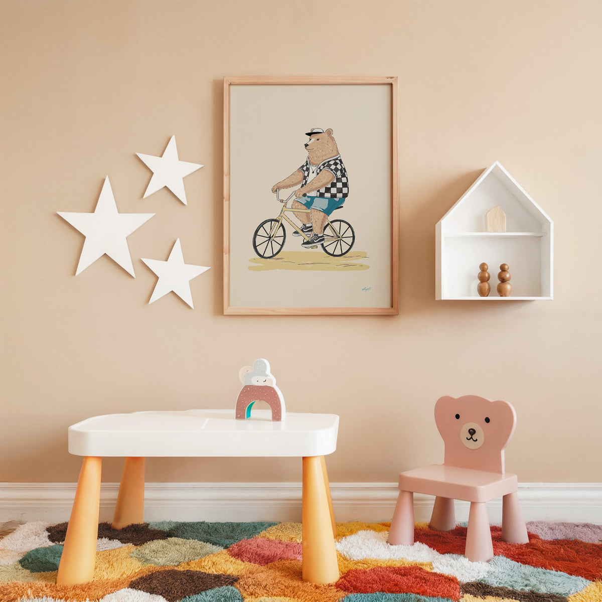 Brown bear riding yellow bicycle nursery print in an oak frame on a peach wall above a kids table with wall stars